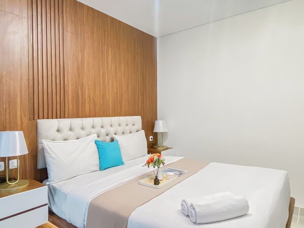 Une chambre d’hôtel avec une tête de lit capitonnée, du linge de lit blanc, un coussin bleu décoratif, une table de chevet avec une lampe et une serviette roulée posée sur le lit.