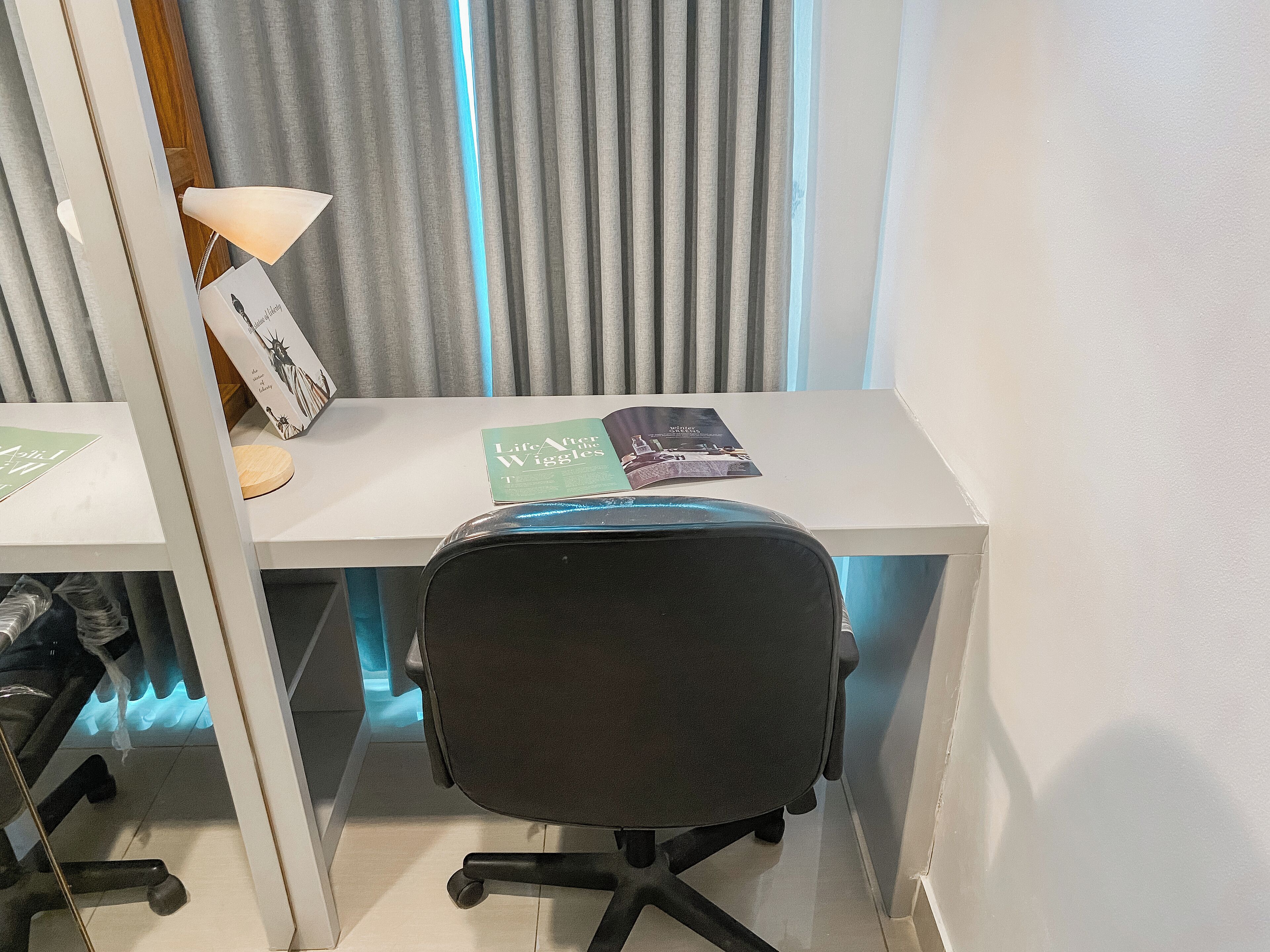 Un espacio de oficina moderno con un escritorio blanco, una silla de oficina negra y una lámpara. Hay dos libros sobre el escritorio, uno titulado 