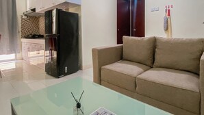 A modern living room with a glass coffee table, beige sofa, and a kitchen area with white cabinets and a black refrigerator. - Homey And Modern 2Br At Marina Ancol Apartment (Jakarta)