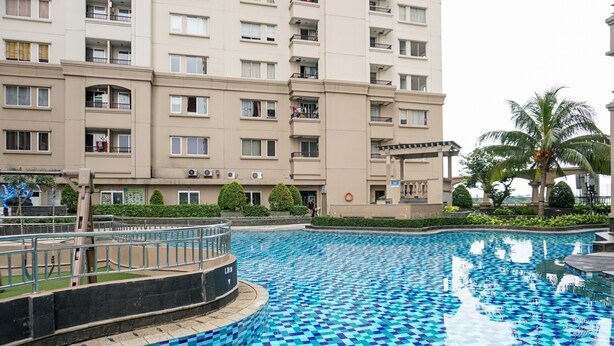 A swimming pool with blue mosaic tiles, surrounded by a low brick wall and a metal railing. In the background, there is a multi-story residential building with balconies and a palm tree. - Homey And Modern 2Br At Marina Ancol Apartment (Jakarta)
