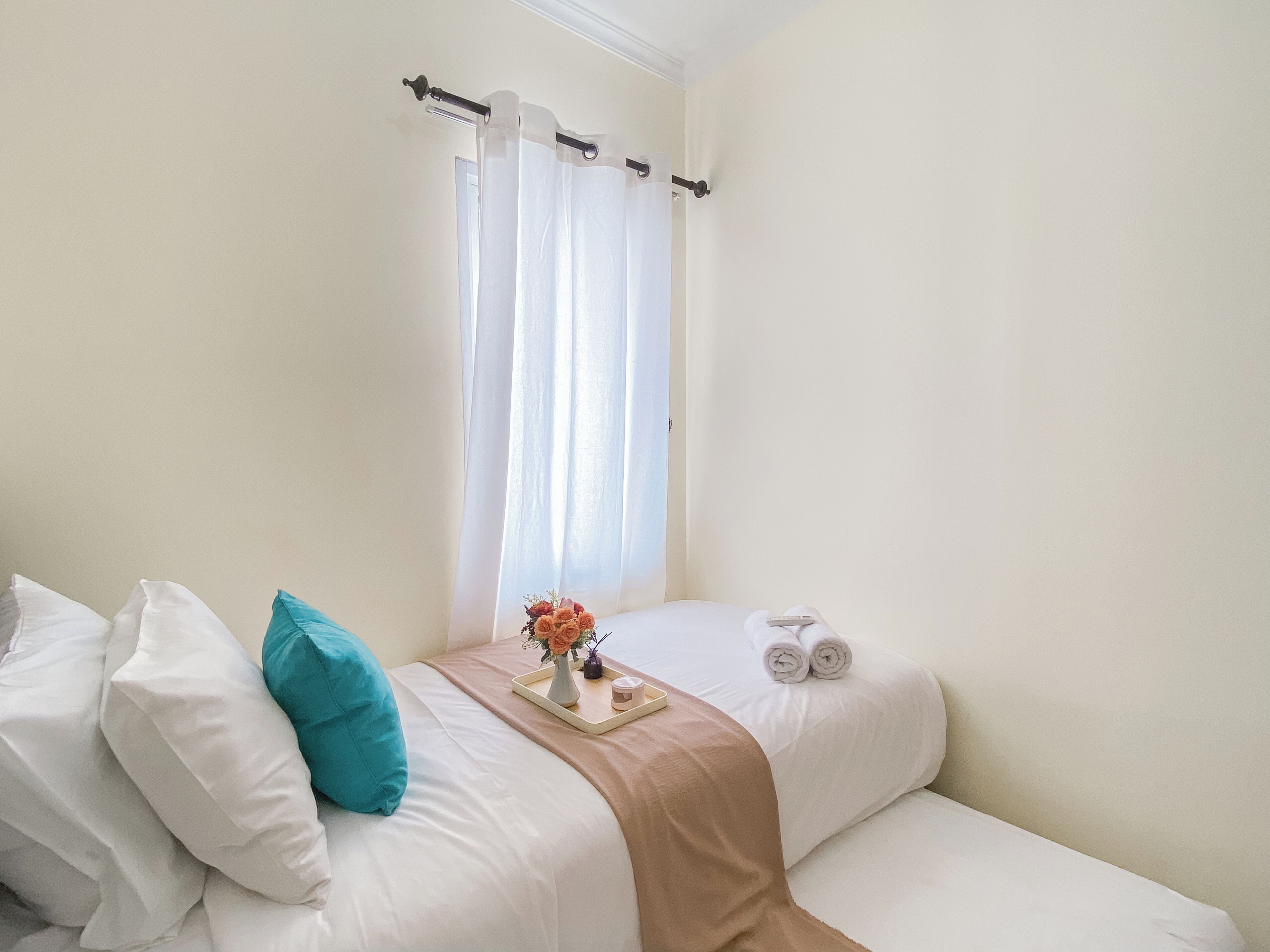 A neatly made bed with white and teal pillows, a beige throw blanket, and a tray with a vase of flowers and a candle.