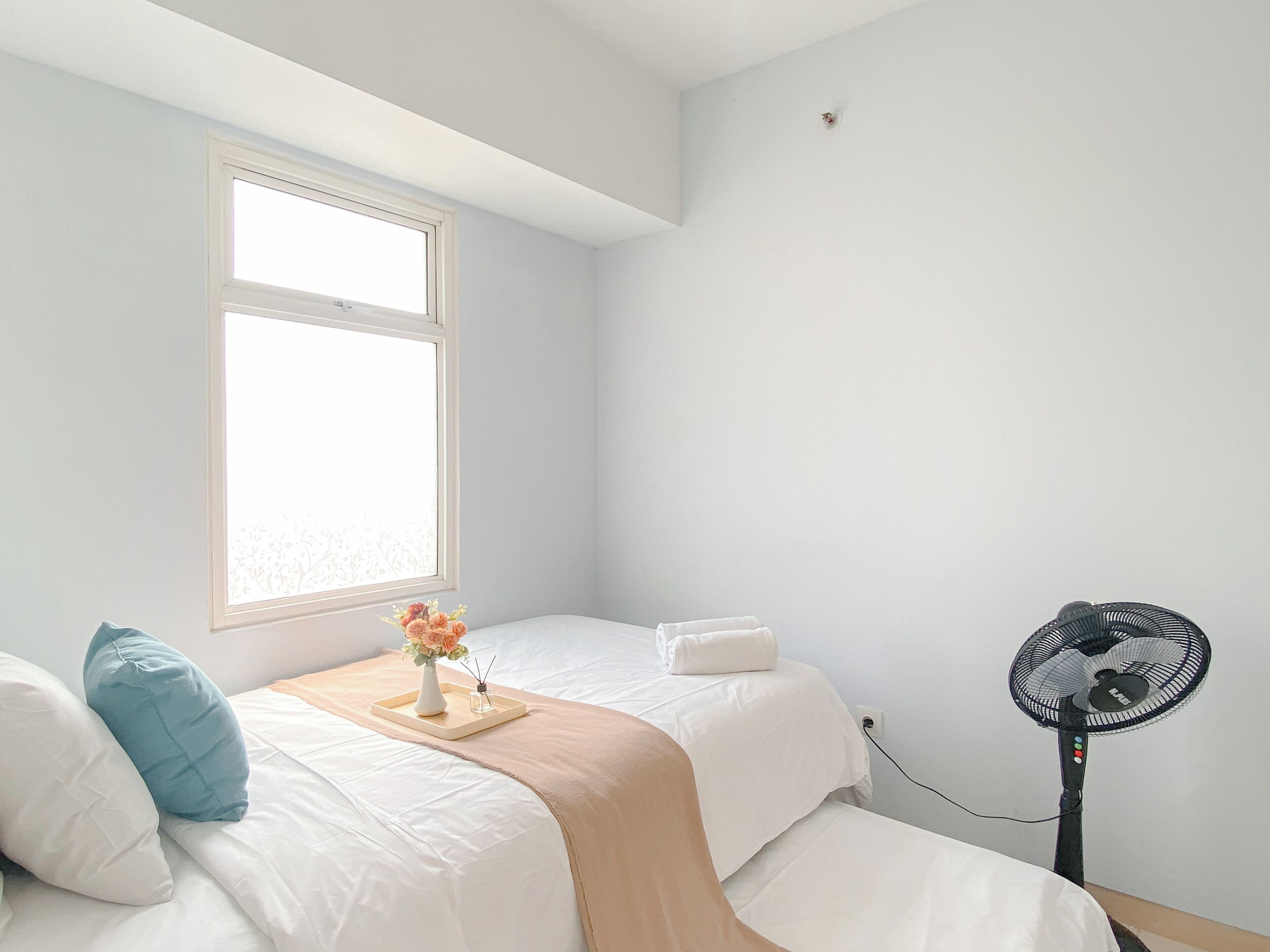 A bedroom with two beds, a window, a bedside table with a vase of flowers, and a standing fan.