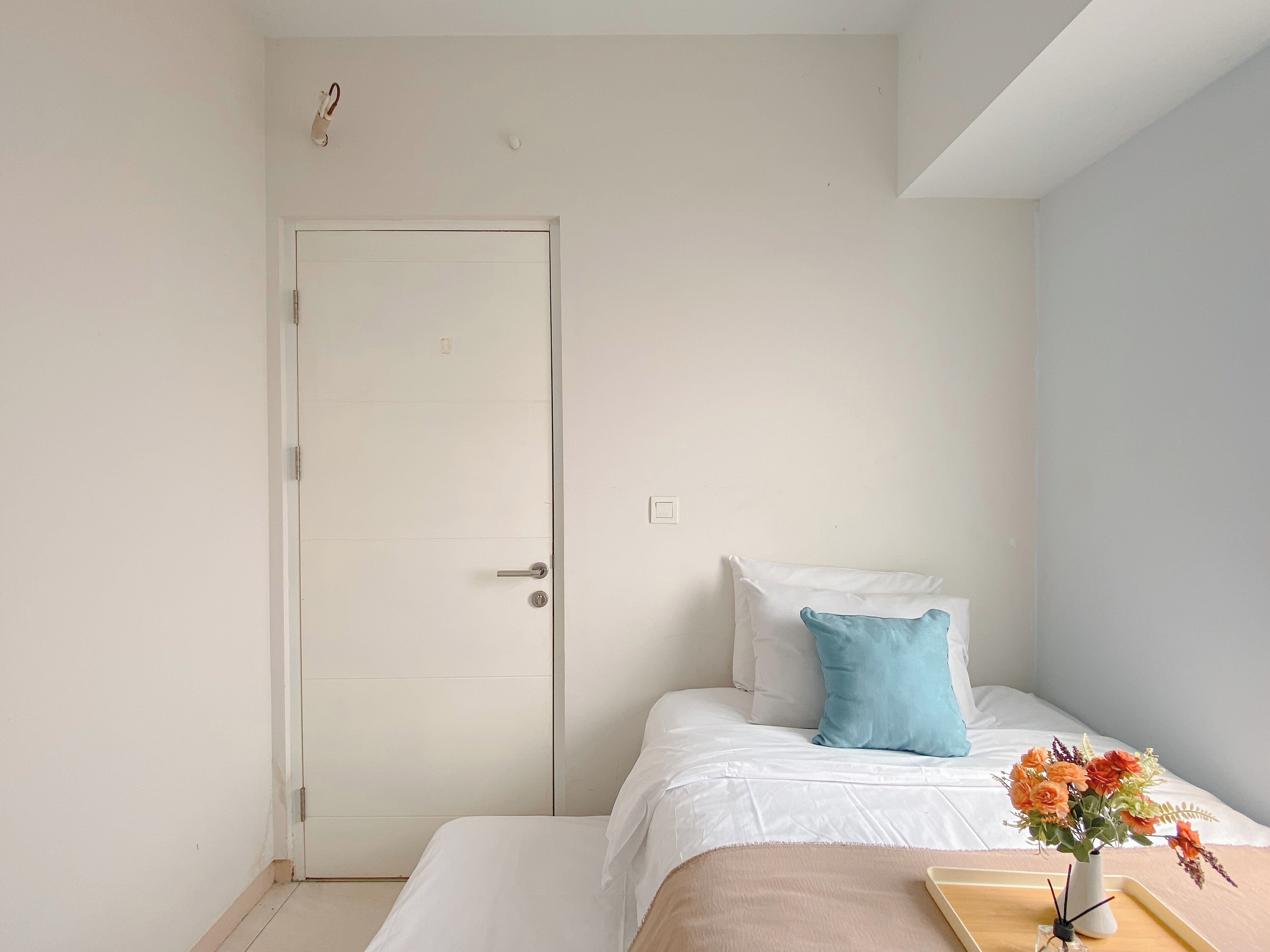 Un dormitorio con una cama, una puerta, una mesita de noche con un florero y un ventanal pequeño.