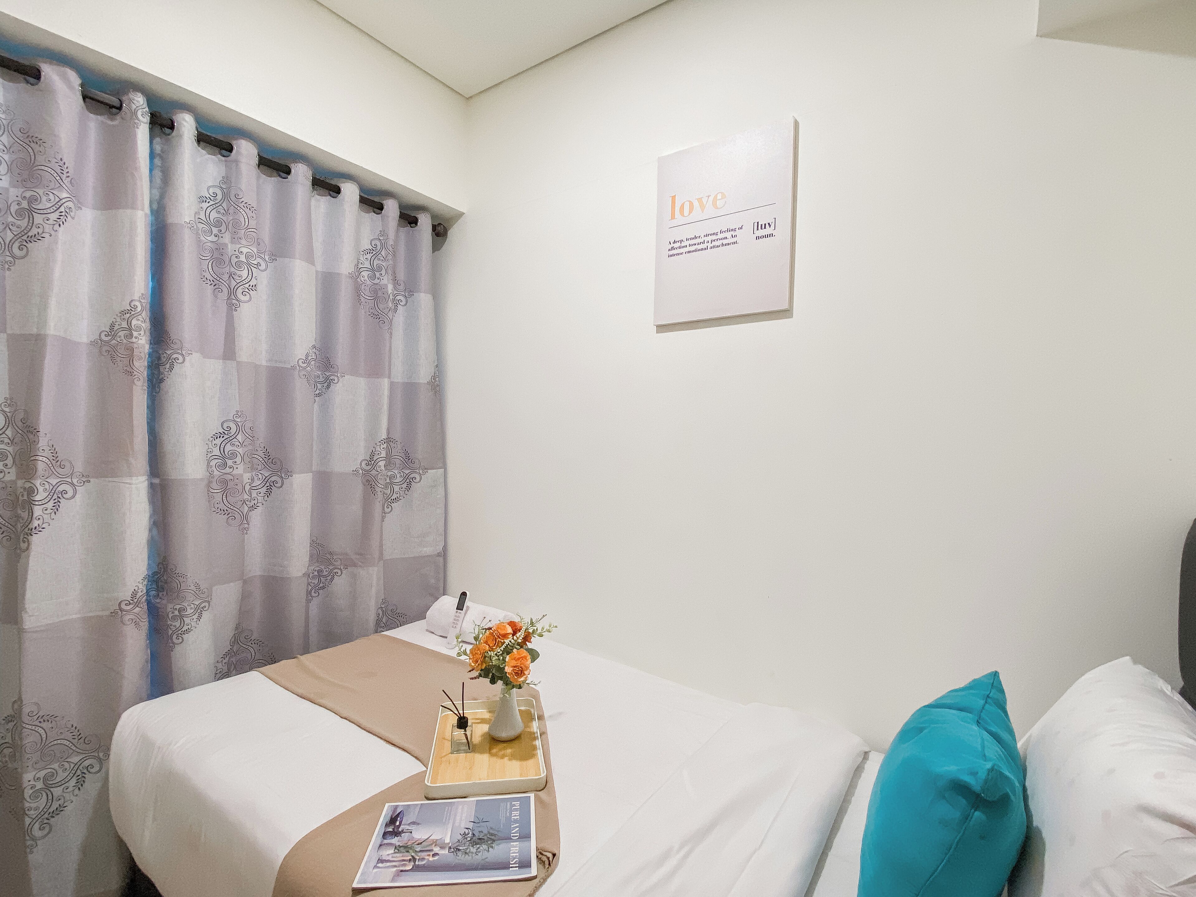Una cama bien hecha con una colcha blanca, un cojín color turquesa y una bandeja de madera con un jarrón de flores y una revista. En la pared, una definición enmarcada de 'amor'.