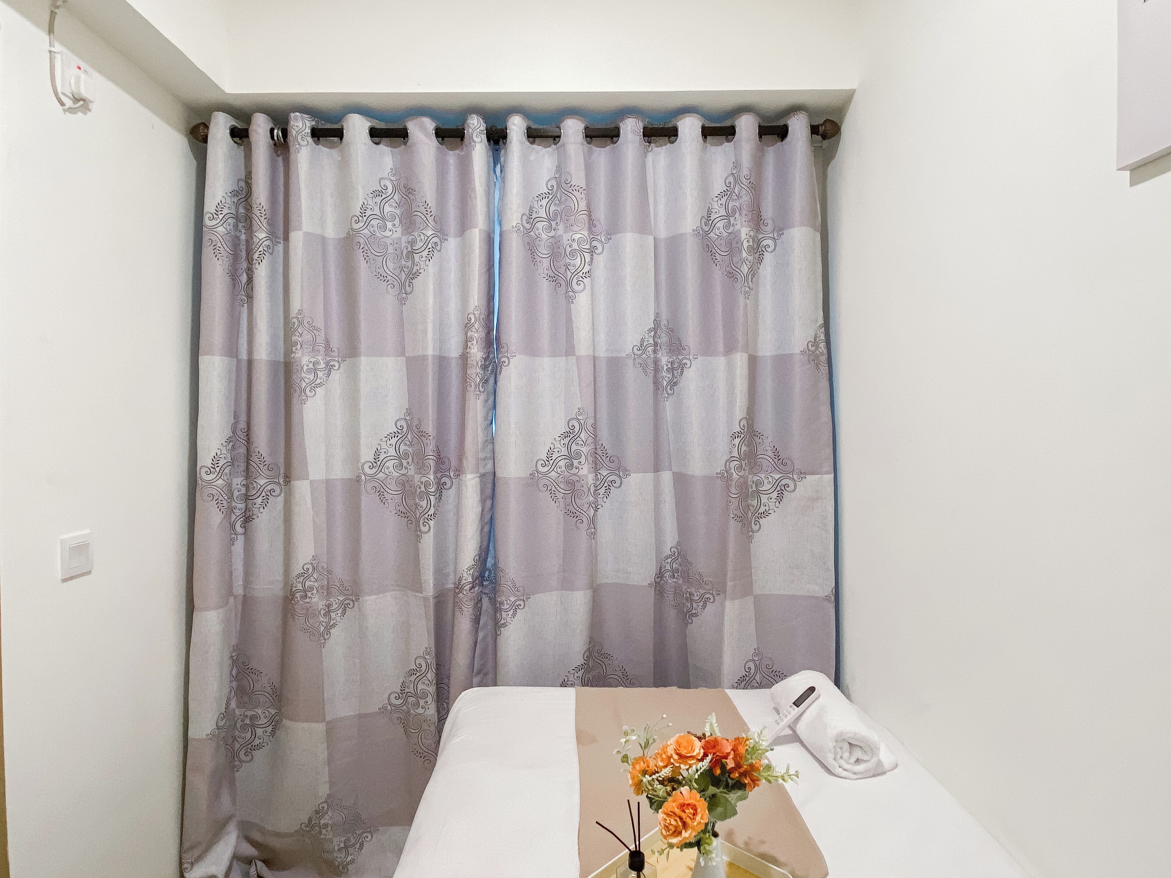 Una habitación de hotel con una cama, una mesita con un florero y un toallón doblado, y una cortina estampada.