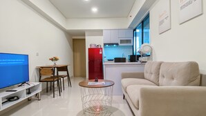 A modern living room with a sofa, a TV, a dining table with chairs, a kitchen area with a refrigerator, and a small side table with a lamp.