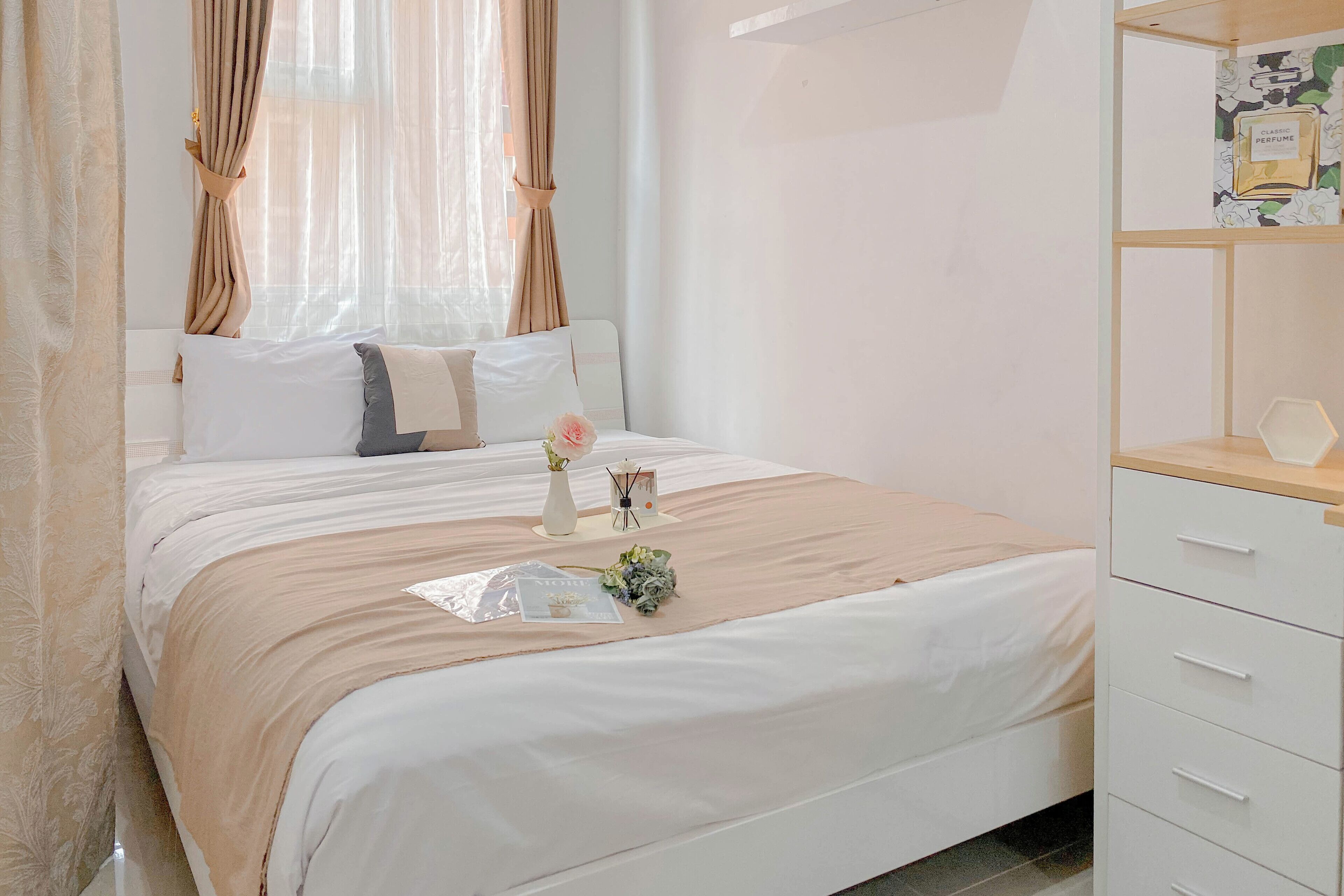 A neatly made bed with a beige bedspread, white pillows, and a small vase with flowers. There is a wooden nightstand with a lamp and a small table with a book and a vase.