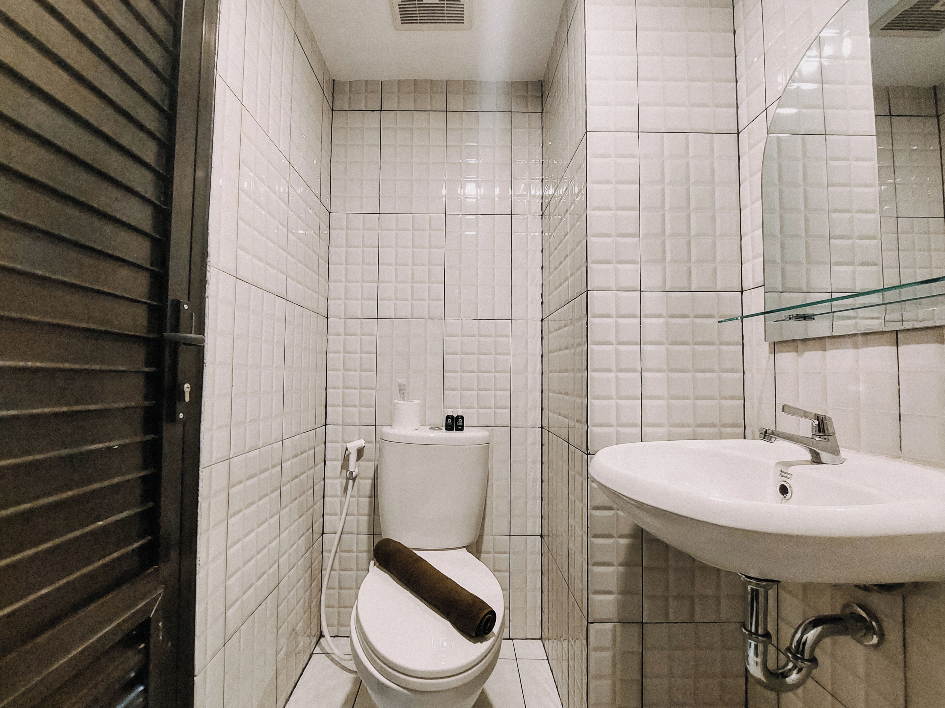 Une salle de bain avec des murs recouverts de carreaux blancs, des toilettes, un lavabo et une étagère en verre.