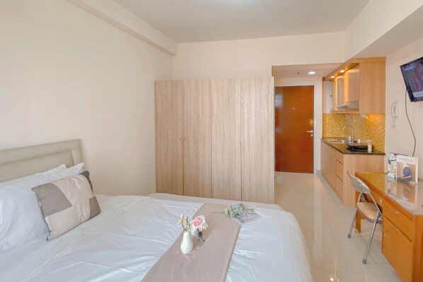 Habitación de hotel con cama, mesita de noche con un jarrón de flores, armario de madera, kitchenette con televisión y baño al fondo.