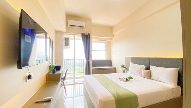 A hotel room with a bed, a TV mounted on the wall, a balcony, and an air conditioning unit. - Comfortable And Homey Studio Room Parkland Avenue Apartment (South Tangerang)
