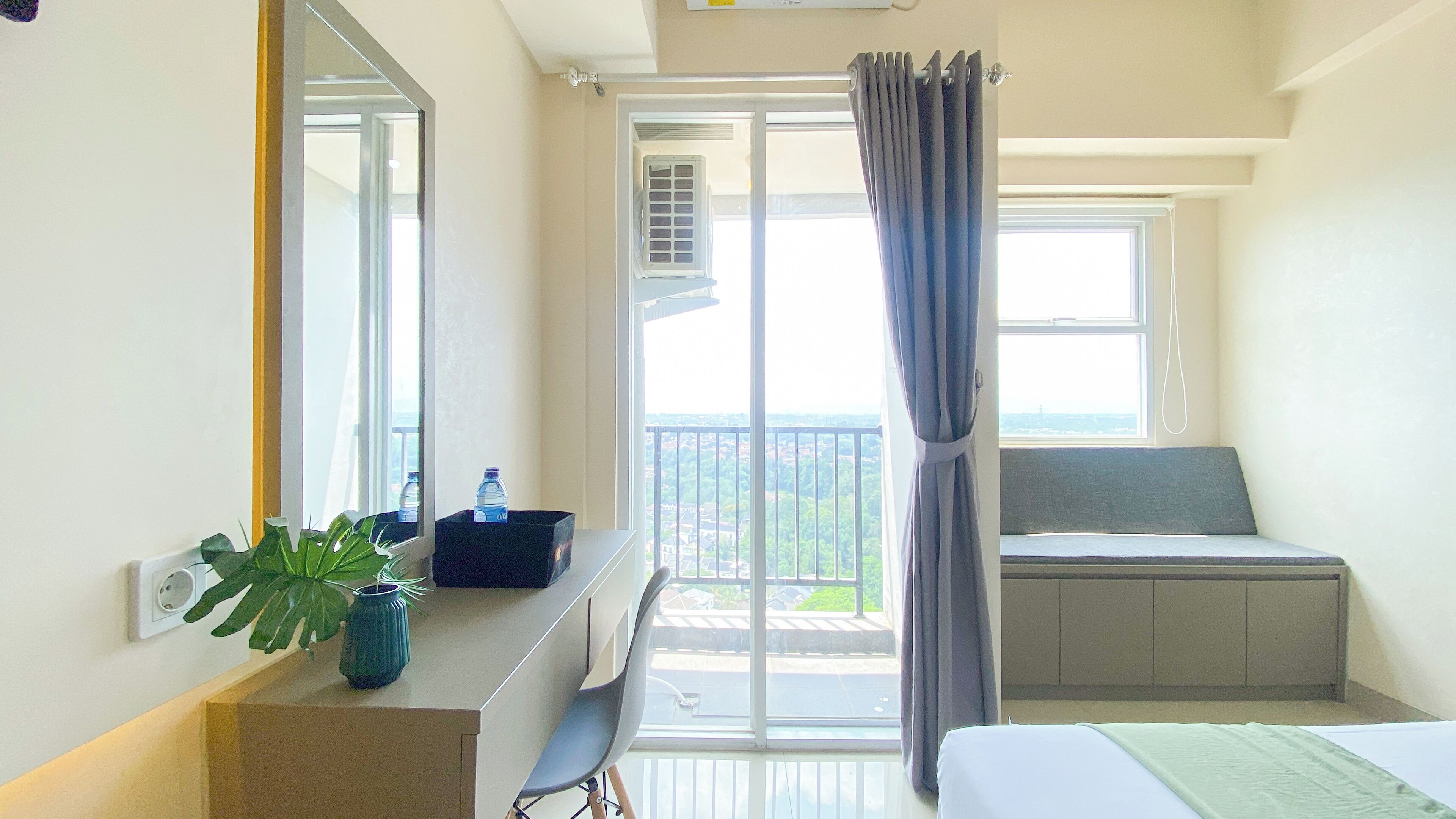 Une chambre d’hôtel comprenant un lit, un bureau avec un miroir, une plante en pot, une fenêtre donnant sur la ville et un balcon avec un siège.