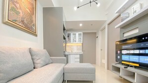 A modern living room with a grey sofa, a flat-screen TV, and a kitchen area in the background. - Cozy 1Br With Extra Room At Vasanta Innopark Apartment (Cikarang)