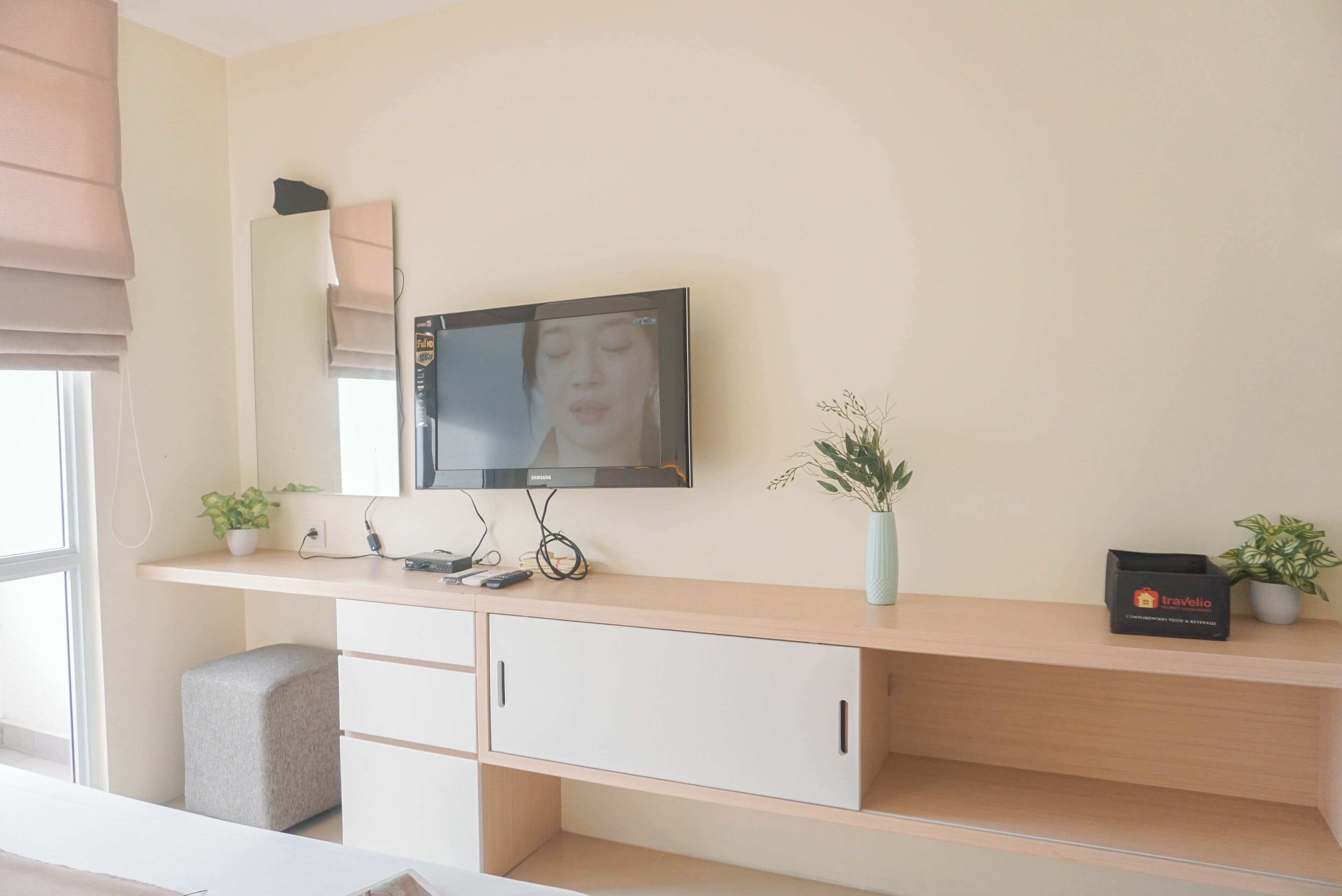 A modern hotel room with a large flat-screen TV mounted on the wall, a wooden console table with a plant, a small seating area, and a travel-themed box on the table.