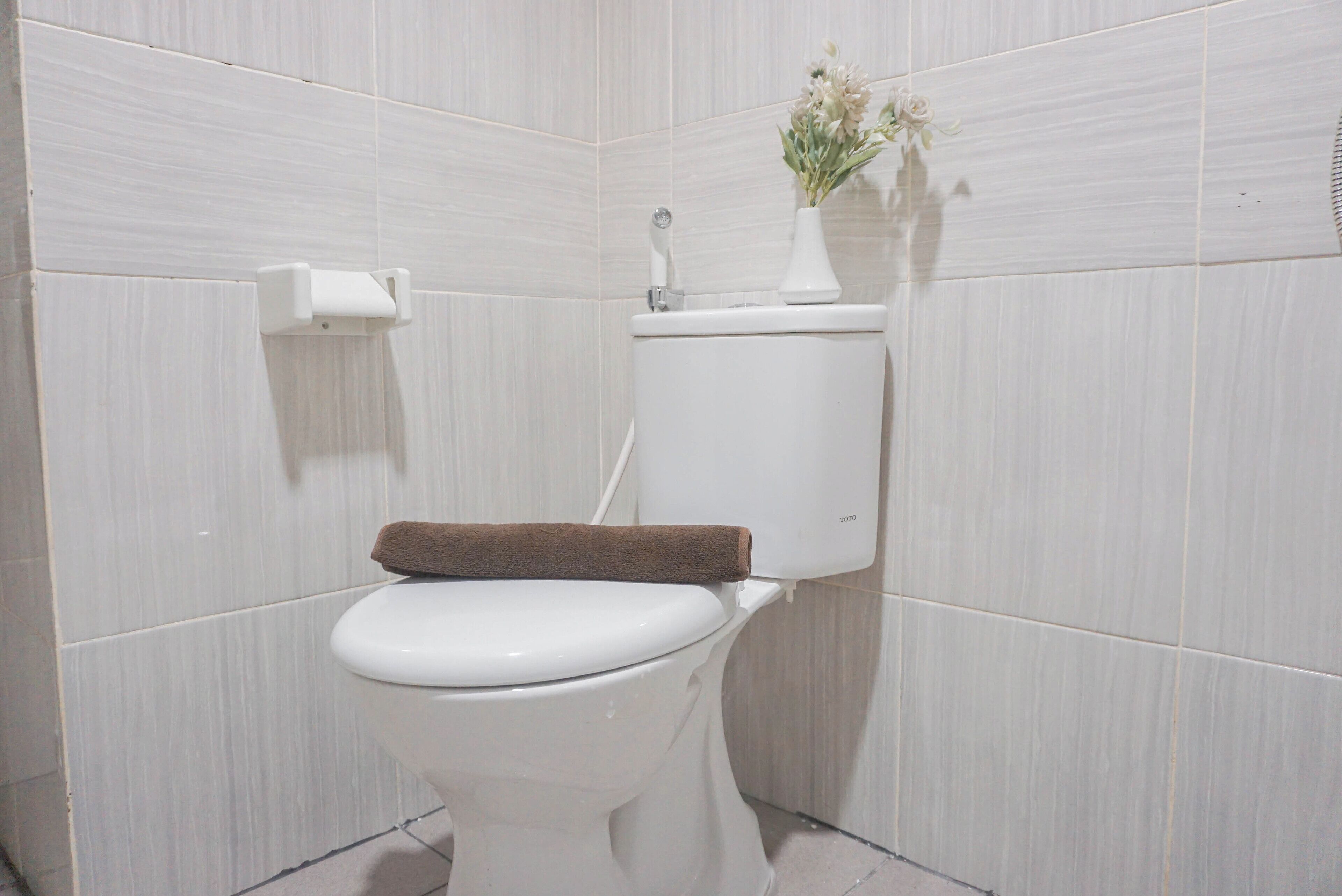 A white toilet with a brown towel on the seat, a toilet paper holder, and a vase with flowers on the tank.