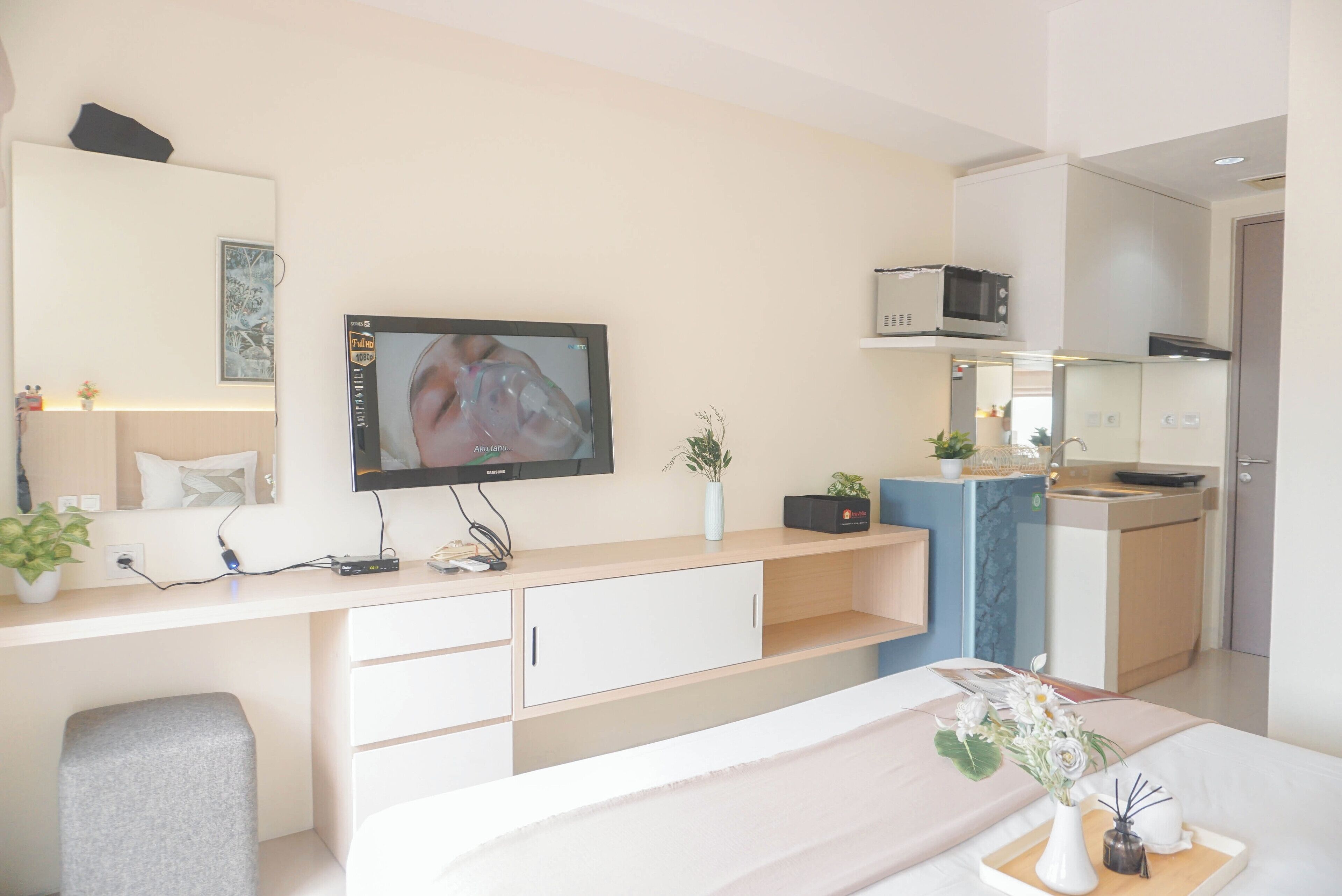 A modern room with a flat-screen TV, a desk with a computer, a chair, a bed, a small kitchen area with a microwave, and a potted plant.
