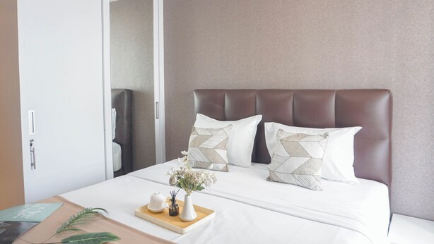 Una cama bien hecha con cabecera marrón, ropa de cama blanca y almohadillas decorativas. Hay una bandeja con un jarrón de flores y una tetera sobre la cama.