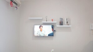 Una habitación con una televisión de pantalla plana en la pared, una cama blanca, una mesita con un florero y dos libros en una estantería.