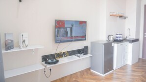 En moderne hotellobby med et fladskærms-tv på væggen, der viser nyhedsudsendelse, en receptionsdisk med kaffemaskine og et lille tekøkken.