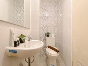 Un baño con un lavabo blanco, un inodoro y un espejo. Hay dos botellas sobre el lavabo y una planta junto al inodoro.
