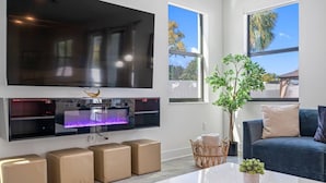 A modern living room with a large flat-screen TV, a built-in fireplace, and a cozy seating area with a blue sofa and cushions. - Luxe Villa • Pool • Spa • Fire Pit • Games & More! (Tampa)