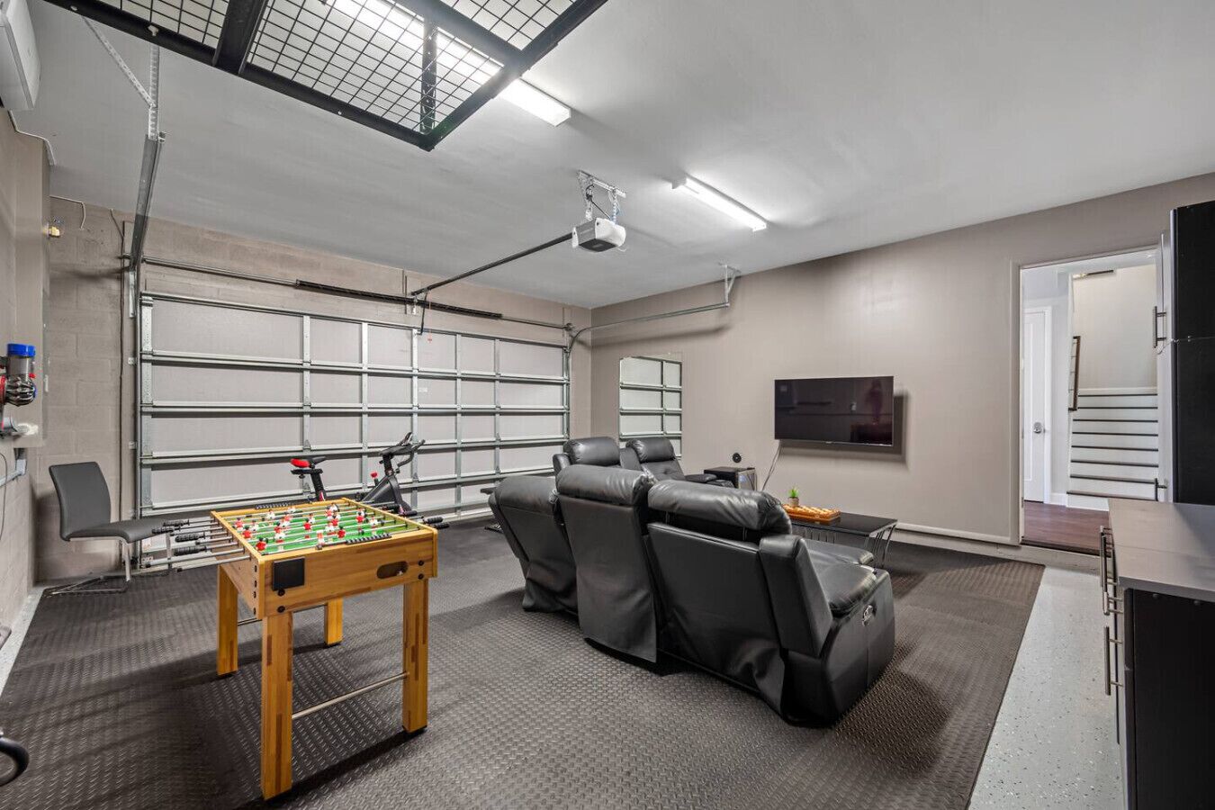 A room with a foosball table, leather chairs, a TV, and a garage door.