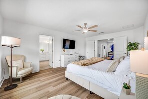 Quarto com cama, cadeira, televisão, ventilador de teto e porta para outro cômodo.