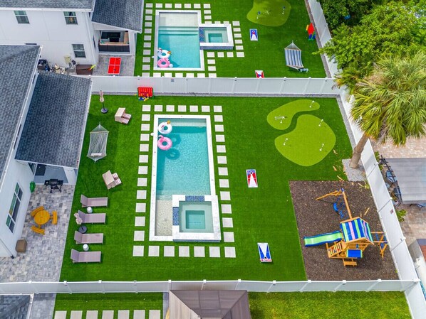 An aerial view of a residential property with two swimming pools, a children's play area, and a patio with lounge chairs.
