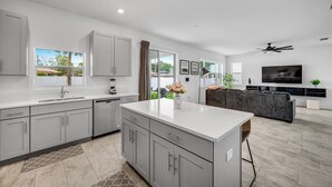 A modern kitchen with grey cabinets, a central island, and a dining area with a dark sofa and a TV. - Family Oasis • Pool • Slide • Games • Sleeps 14+ (Tampa)