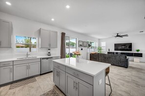 A modern kitchen with grey cabinets, a central island, and a dining area with a dark sofa and a TV.