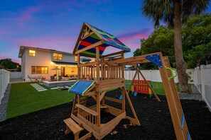 Estructura de madera para juegos con un tobogán y columpios, frente a una casa y un cielo crepuscular.