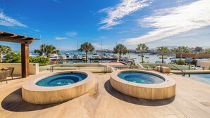 Zona de piscina en la azotea con dos jacuzzis, con vistas a un puerto deportivo con barcos y un cielo azul despejado.