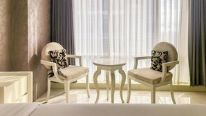 Una habitación con dos sillas blancas con cojines estampados, una mesita redonda y cortinas de encaje en las ventanas.
