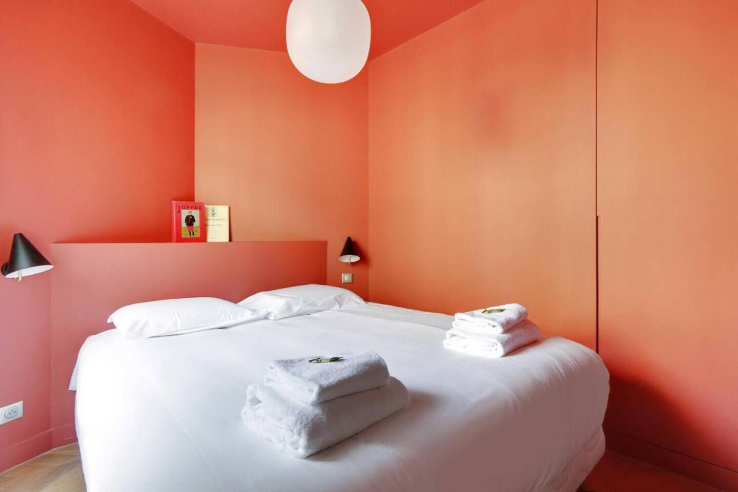 A hotel room with a large bed, white linens, two bedside lamps, a red wall, and a white ceiling light.