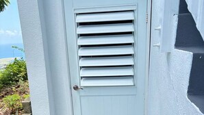 A white door with horizontal slats, a black doormat, and a concrete step leading to a tiled entrance. - Sleek & Sunny Island Studio | Kitchenette (St. Thomas)