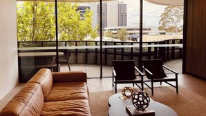 Une pièce moderne avec un canapé en cuir, deux fauteuils noirs, une table ronde et un vase décoratif. De grandes fenêtres offrent une vue sur les bâtiments et les arbres.
