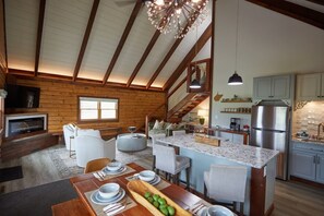 Amplia sala de estar con techo alto, vigas de madera, chimenea y una mesa de comedor lista con platos y un cuenco de fruta.