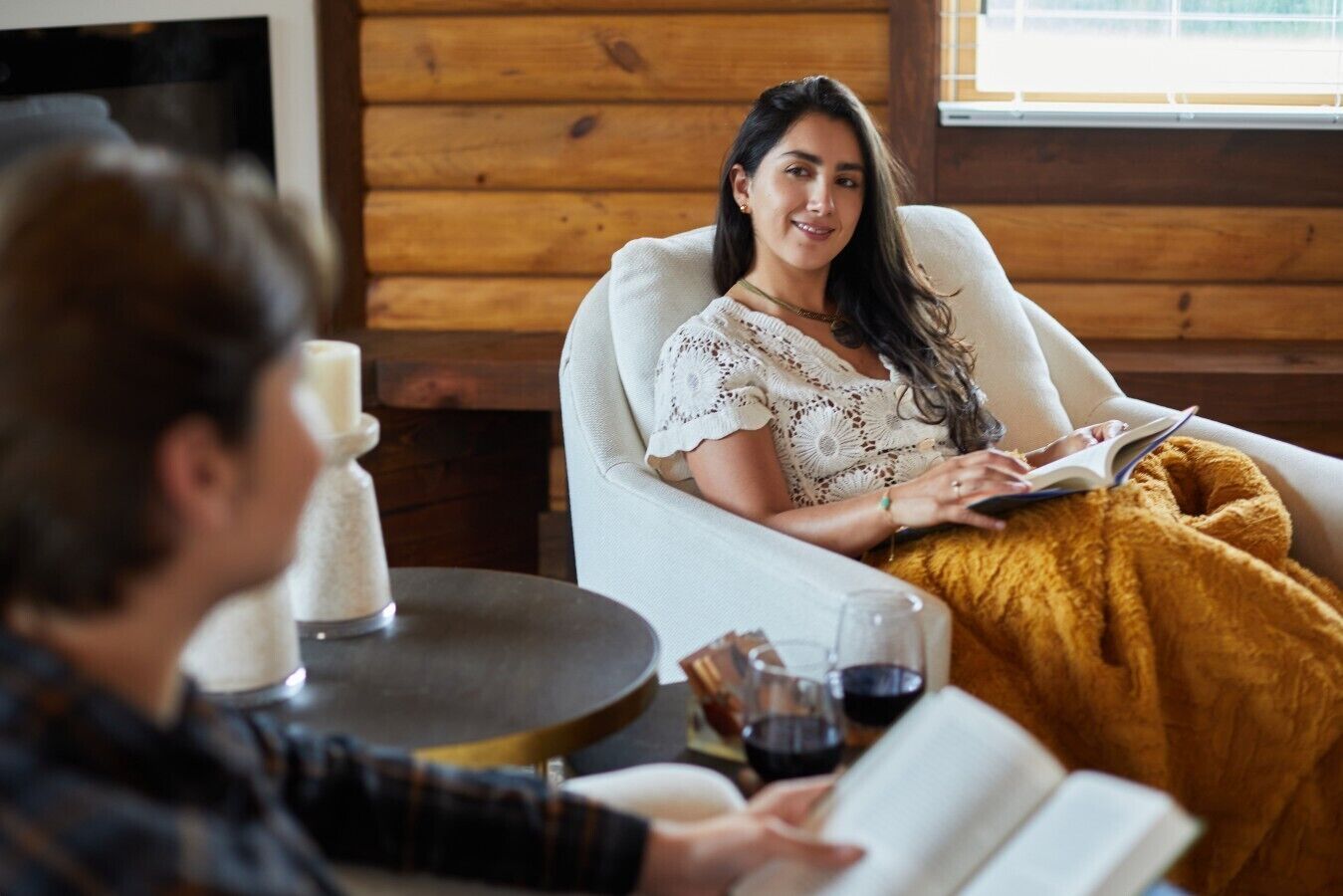 A person sitting in a chair with a book, another person holding a book, and a table with a candle and glasses.