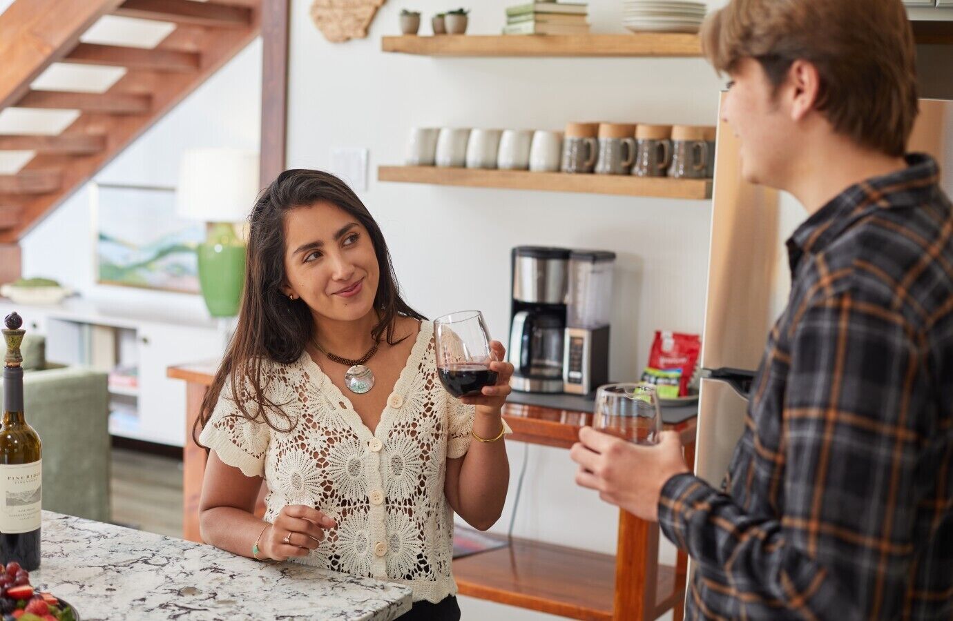 A person holding a wine glass in a room with a coffee maker and shelves with mugs.