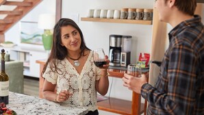 Eine Person hält ein Weinglas in einem Raum mit einer Kaffeemaschine und Regalen voller Tassen.