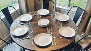 A round dining table set for eight with black chairs, a wooden table, and a view of the outdoors through large windows. - Home Sweet Home (Sutherlin)
