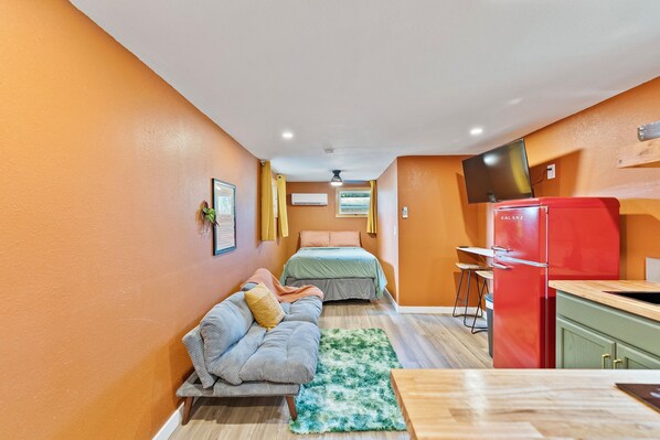 A compact room with a sofa, a bed, a small kitchen area with a red refrigerator, and a TV mounted on the wall.