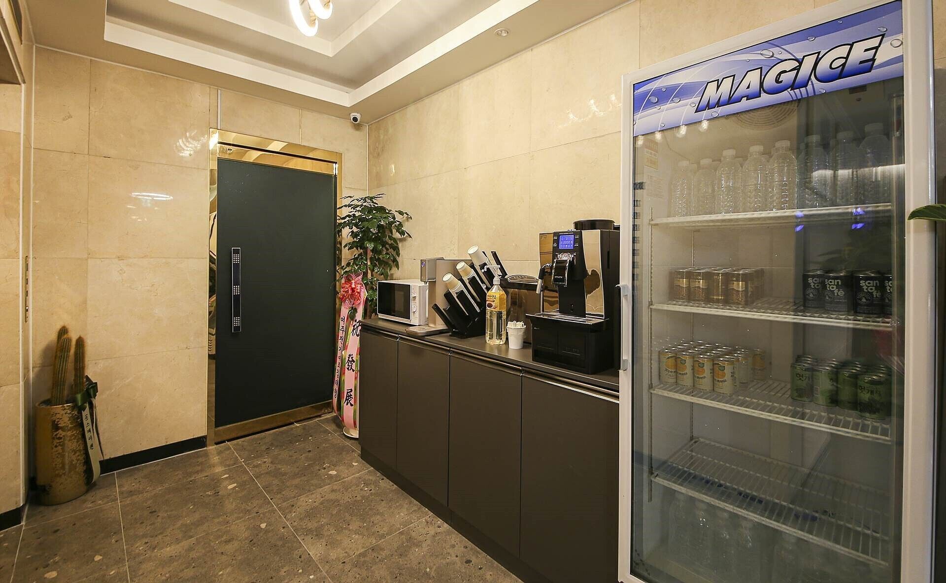 A reception area with a counter, a coffee machine, a refrigerator stocked with beverages, and a door with a security camera.