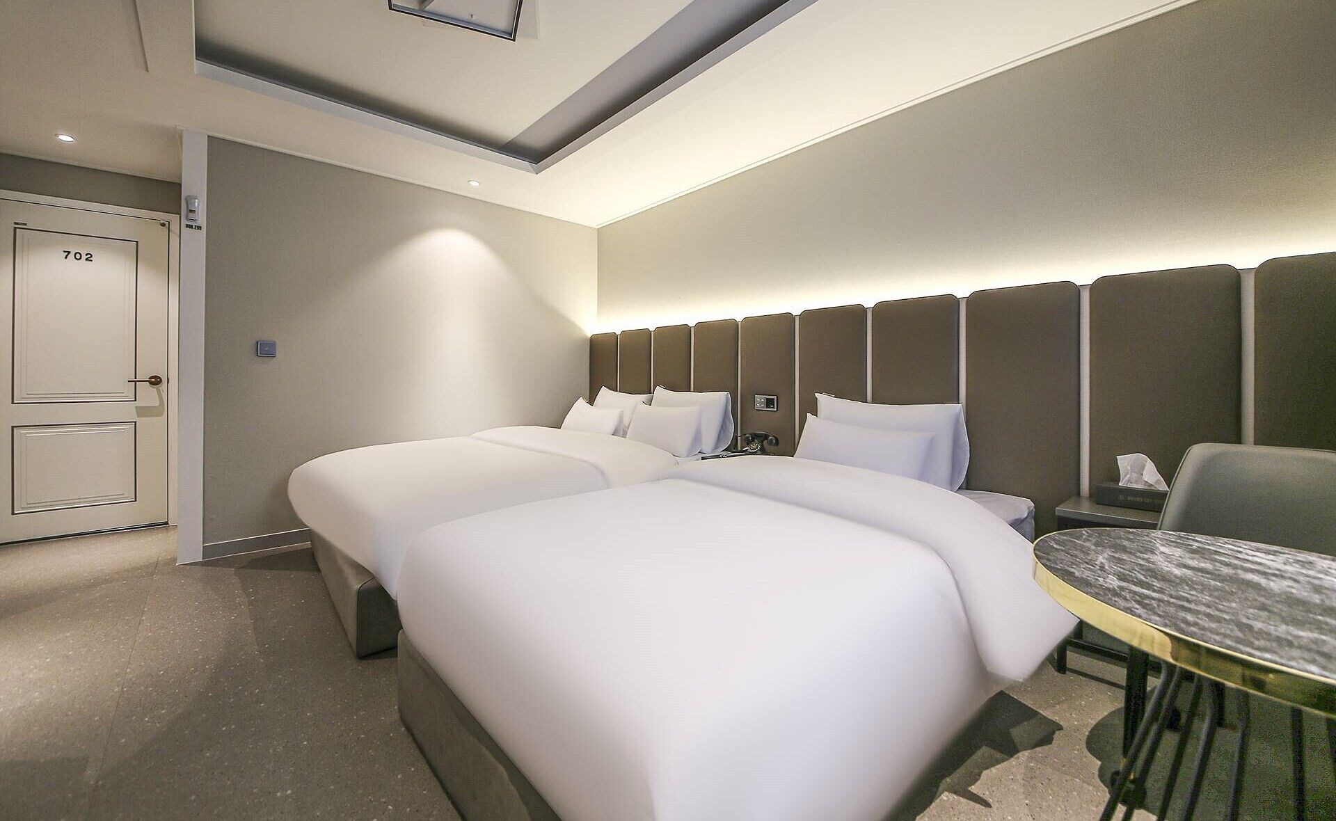 A hotel room with a double bed, a desk with a chair, and a door marked with the number 702.