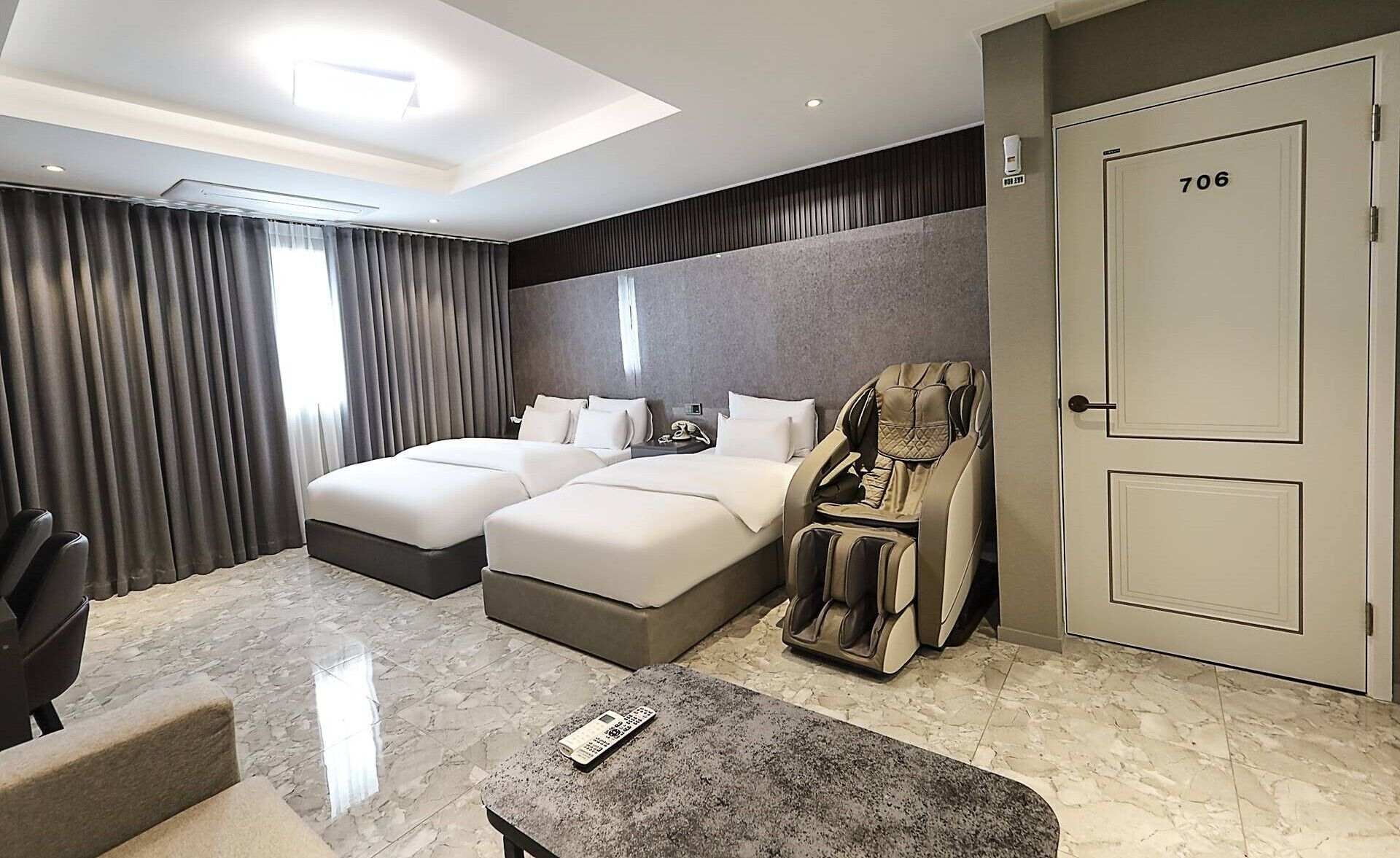 A hotel room with two beds, a massage chair, a desk with a remote control, and a door marked 706.