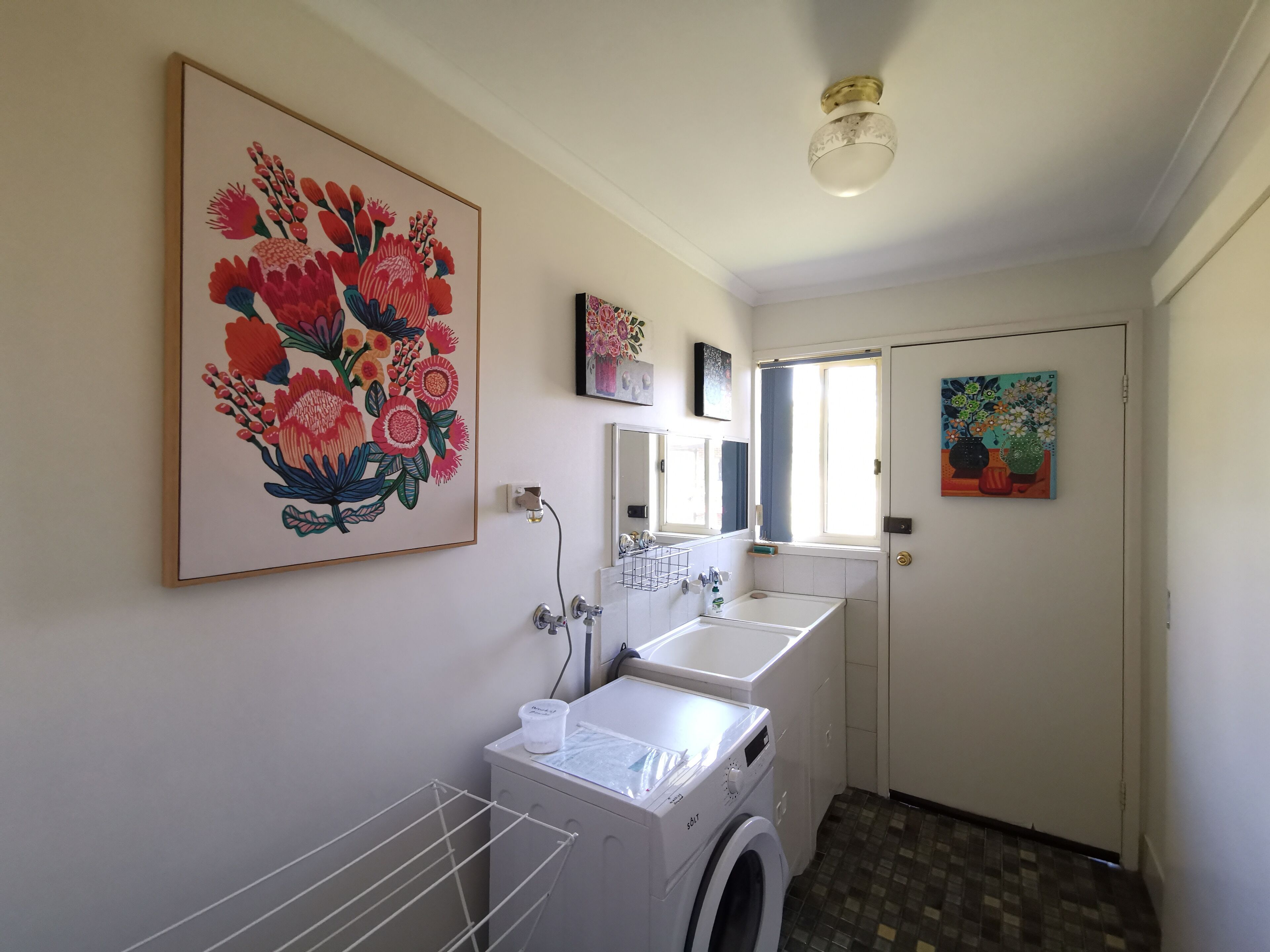 Une buanderie équipée d’un lave-linge, d’un évier et d’un étendoir. Les murs sont décorés de tableaux représentant des motifs floraux.