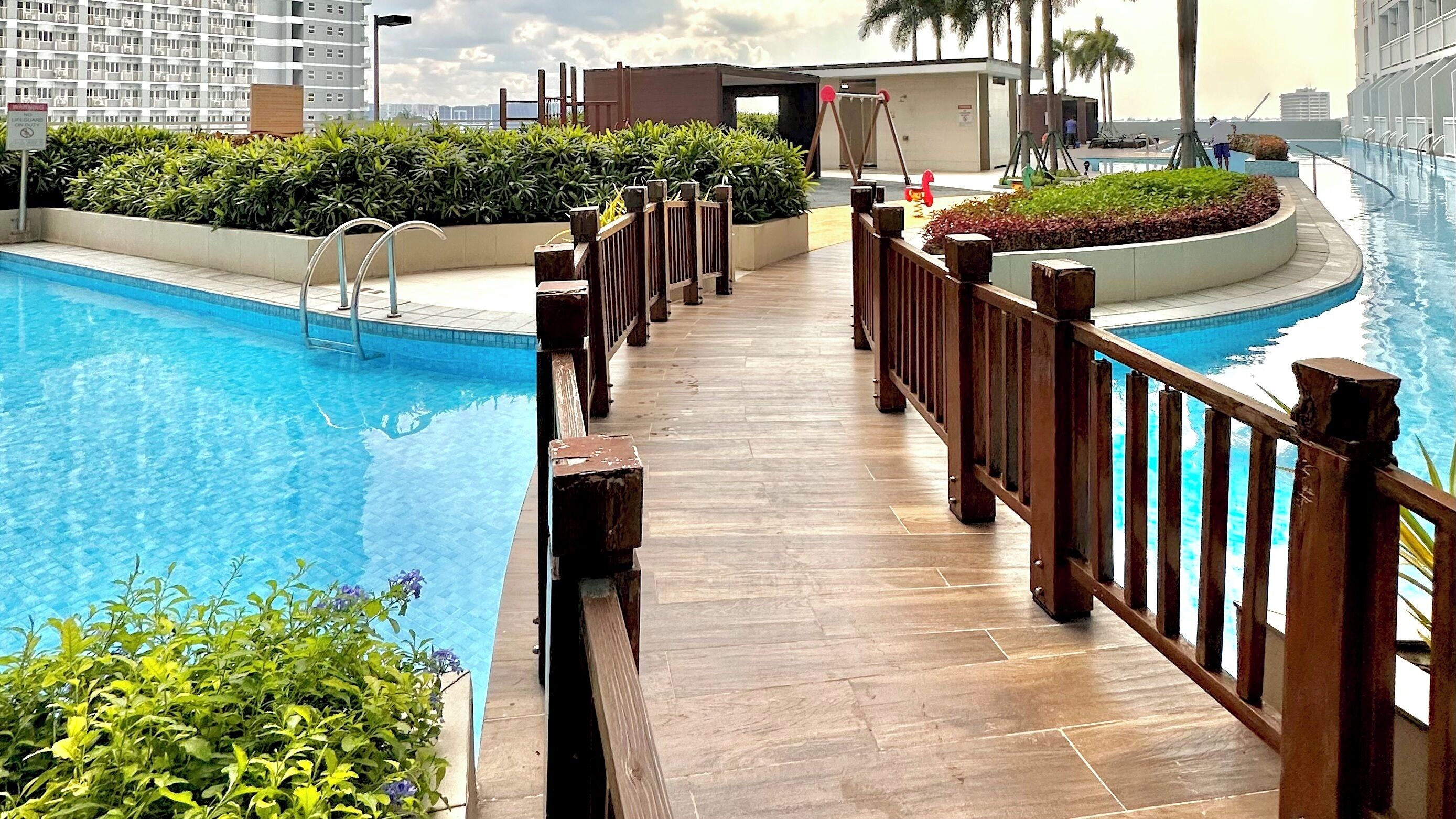 A swimming pool with wooden deck and railings, surrounded by greenery and modern buildings.