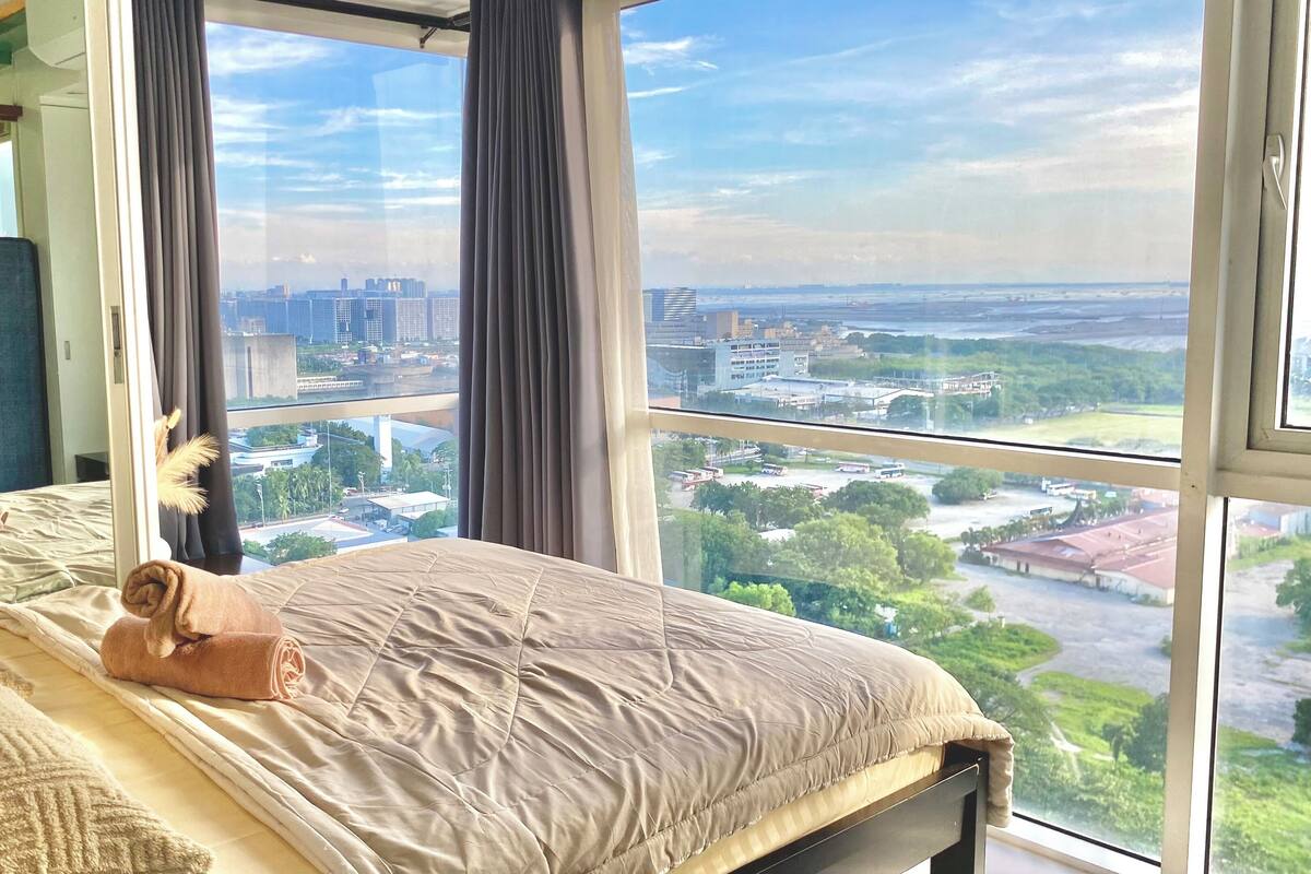 A hotel room with a large window offering a city view, a neatly made bed with a rolled towel, and a blue headboard.