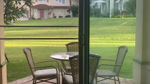 A balcony with wicker chairs and a table, overlooking a well-maintained lawn and a building with a red roof.