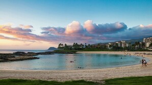 A beach with clear waters, a rocky shoreline, and a resort area with buildings in the background.