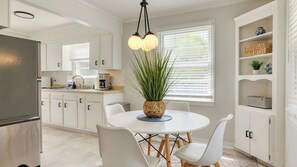 Une cuisine moderne avec des meubles blancs, une table de salle à manger ronde entourée de chaises, une machine à café et une plante en pot.